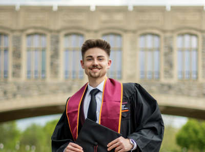 Photo of Giovanni Heater holding graduation cap in front of Torgersen Bridge